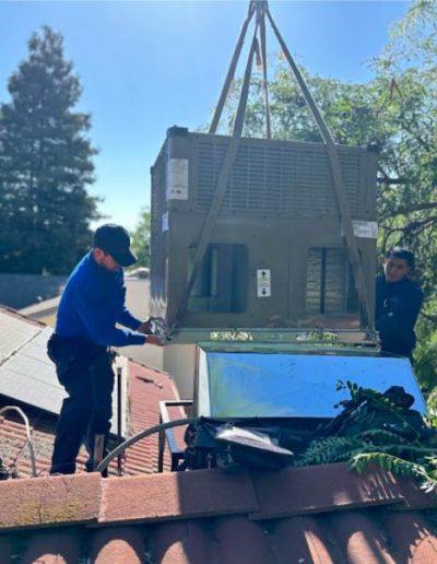 Two Mechanical Air technicians carefully lowering a large, dark green residential HVAC unit onto a red tile roof with straps, surrounded by green trees and with solar panels visible on an adjacent roof under a bright sky.
