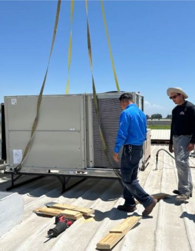 HVAC technicians from Mechanical Air installing a large commercial rooftop air conditioning unit with yellow straps, a sunny day, with two technicians observing and one walking towards the unit, on a corrugated metal roof