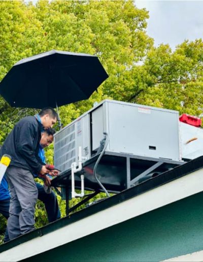 Mechanical Air technicians installing a residential rooftop HVAC unit on a sloped roof under an umbrella on a cloudy day, with a ladder propped against the house and green trees in the background