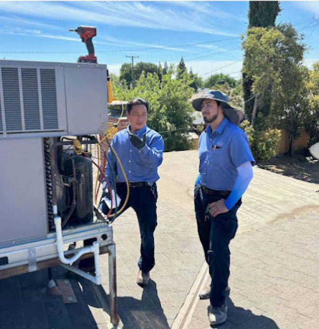 Two technicians are standing on a roof discussing the job next to an open AC unit.
