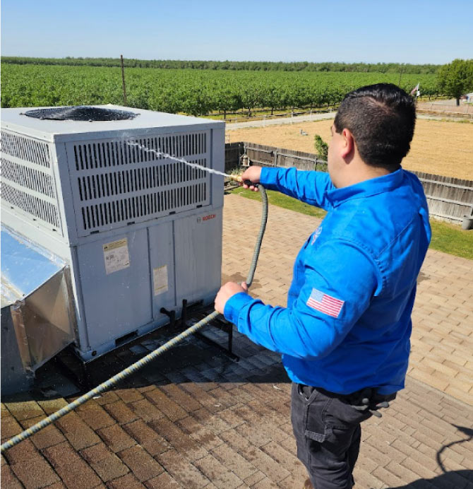 Mechanical Air technician performing rooftop HVAC maintenance by washing down a Bosch package unit with a water hose in a Central Valley agricultural area