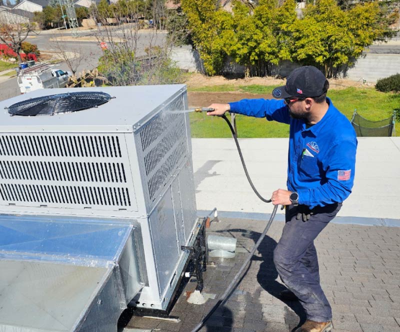 Professional HVAC technician in a blue uniform power washing a commercial rooftop air conditioning unit to clear debris and optimize cooling efficiency, for ac repair in kingsburg, ca